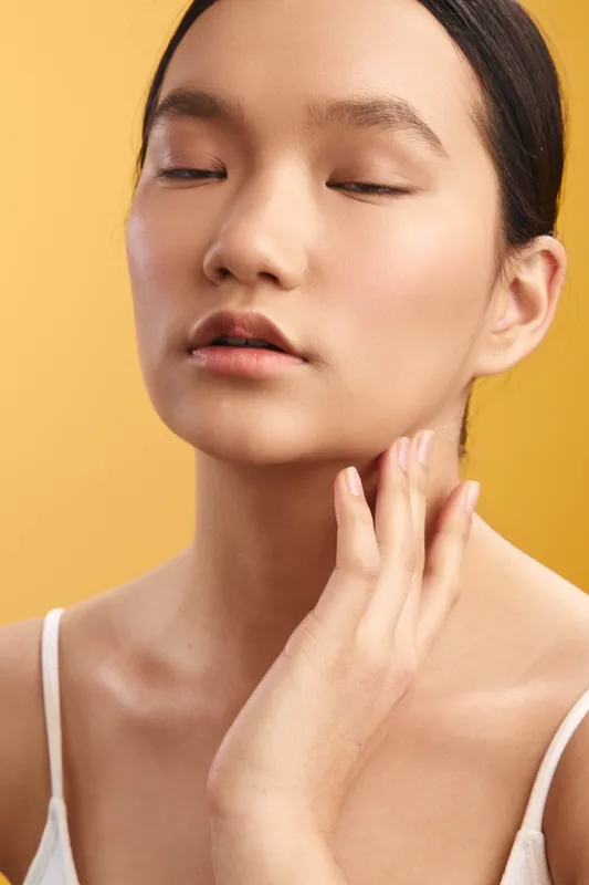 Asian woman closes her eyes in a serene portrait, touching her face with gentle fingers, conveying calm, beauty, and wellness. Soft studio light emphasizes smooth skin and a minimalist mood.