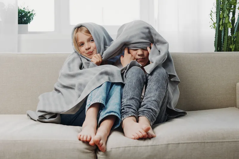 Two children wrapped in gray blanket sitting barefoot on couch, playing and hiding faces in bright living room, cozy home atmosphere.
