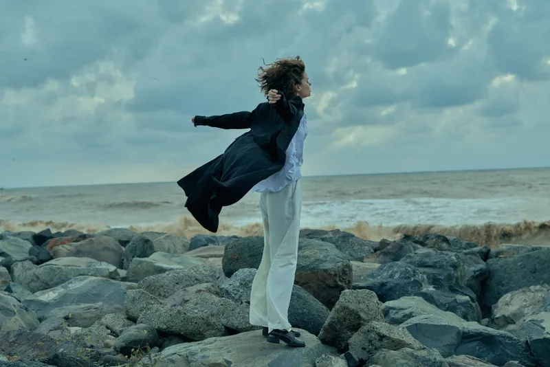 Woman balancing on a rocky shoreline wearing a flowing coat, wind whipping hair, dramatic sea scene conveying freedom, exploration and resilience by the coast