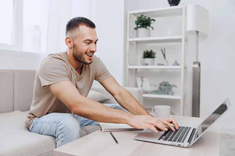 Smiling man engaged in remote work from home, gracefully typing on laptop, showcasing modern lifestyle and comfort in a cozy, bright living space.