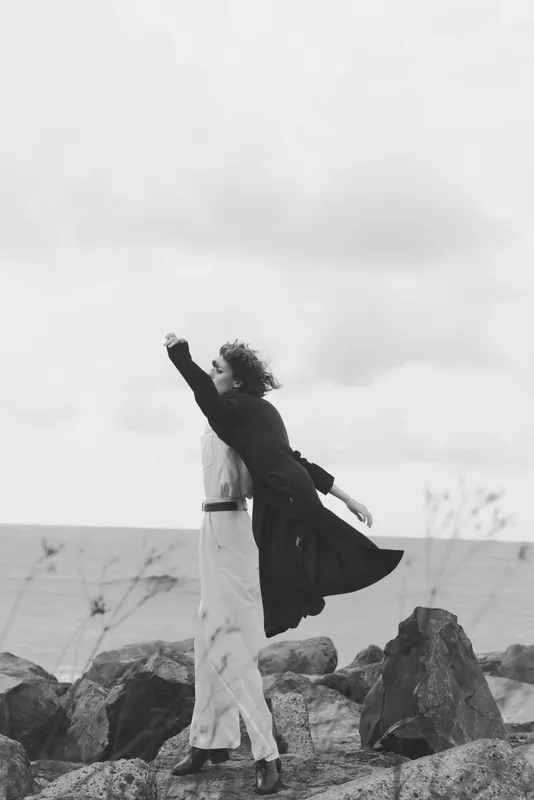 Dancing couple on a rocky seaside, silhouettes meet the wind as coats swirl and the horizon glows in moody black and white, an artistic lifestyle moment