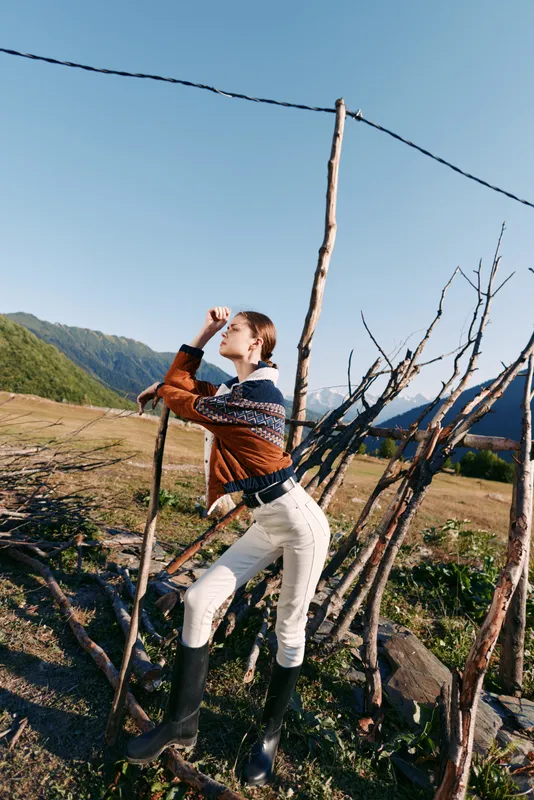 Woman in countryside leaning on rustic fence wearing patterned sweater and tall boots, fashion portrait with mountains background, natural light, rural style and contemplative pose.
