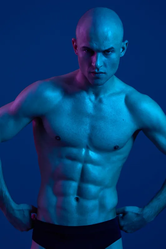 Strong man posing confidently against a vibrant blue background, showcasing his muscular physique in a unique light. A striking and evocative black and white image.