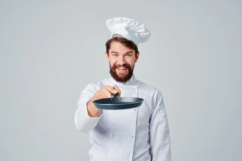 Мужчина в поварской форме с кастрюлей готовит на кухне на светлом фоне