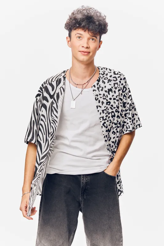 young man wearing a multipattern leopard and zebra print shirt with casual white t-shirt and black jeans standing with hand in pocket against white background