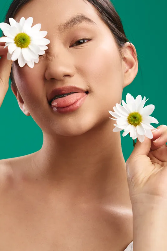 Asian woman is featured in a closeup portrait with flowers, showcasing fresh beauty and playful charm. A bright studio setting complements natural makeup and a serene expression.