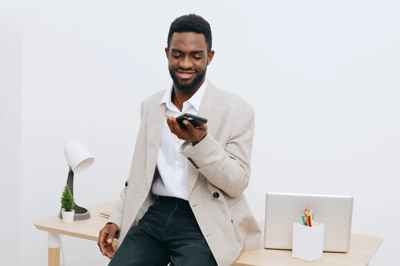 Smiling man using mobile phone while standing at modern office desk, casual business attire, enjoying technology, concept of communication, remote work, digital lifestyle, indoors on neutral backgroun