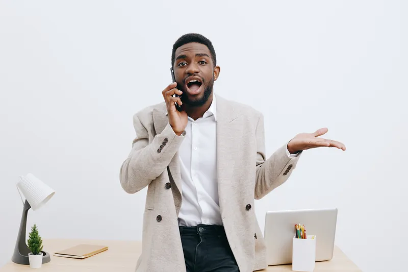 Confident smiling man talking on mobile phone indoors at workplace, casual business attire, energetic expression, modern office background, white wall, professional atmosphere, digital communication c