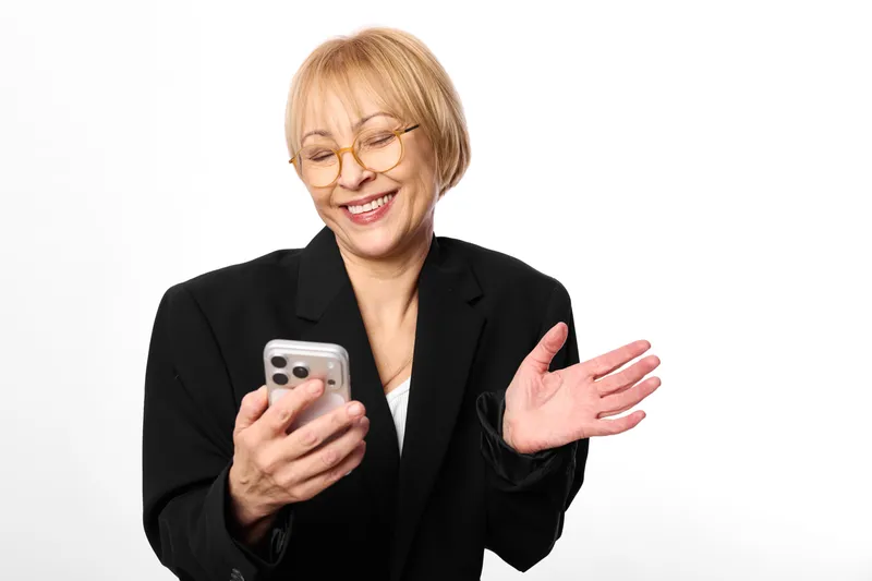 Smiling senior woman in black blazer holds smartphone, waves her hand, cheerful business communication concept, isolated on white background, copy space portrait