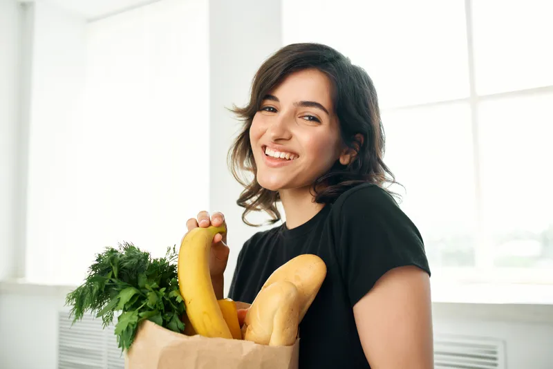 Весёлая женщина с пакетом продуктов занимается домашними делами, связанными со здоровой пищей