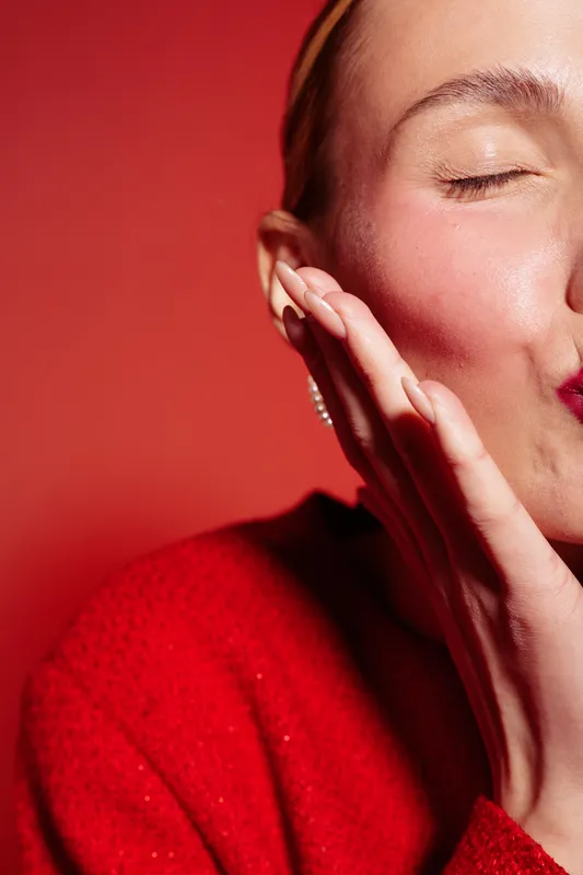 Close-up of a woman with closed eyes touching her cheek, wearing a red sweater and headband. The warm red background creates an intimate and cozy mood for skincare or beauty concept.