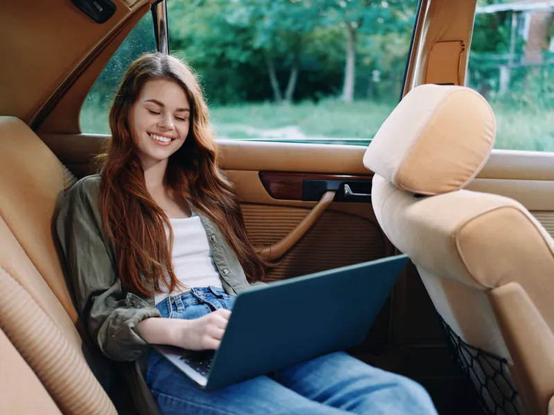 Young woman working on a laptop in a vintage car, smiling and enjoying a peaceful moment in nature, highlighting creativity and leisure.