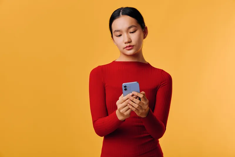 Young woman in red sweater looking at phone for texting or social media communication on orange background. Casual lifestyle and focus on mobile device with relaxed posture indoors.