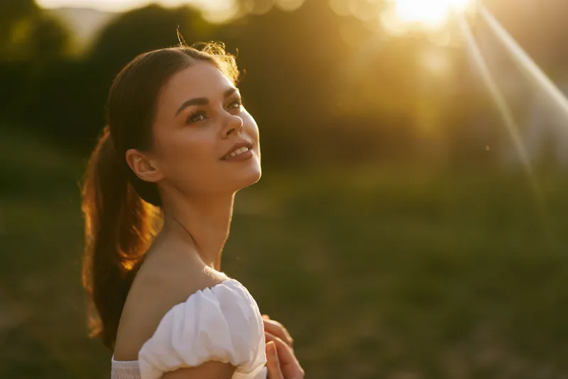Young woman looking up with a serene expression during sunset, wearing a white dress in a natural outdoor setting, evoking feelings of peace and joy