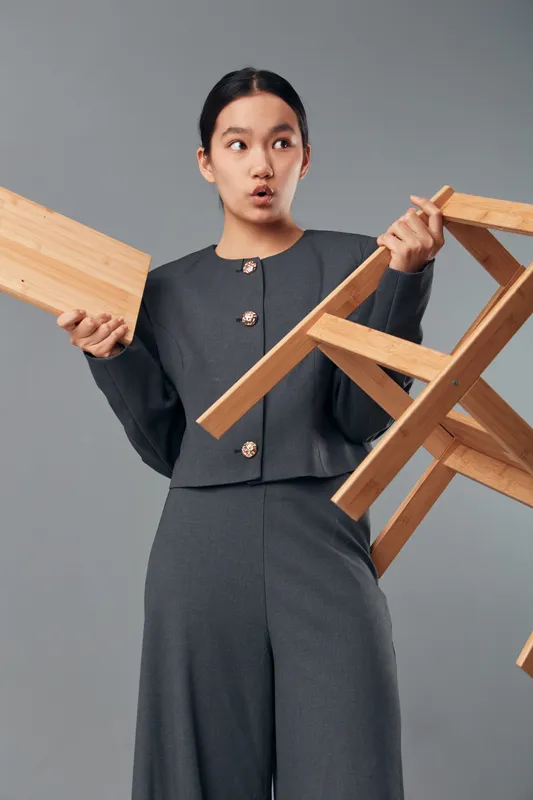 Woman dressed in gray outfit holds a wooden chair and a wooden plank with surprised expression on neutral background. Studio portrait with minimalistic style and casual pose.