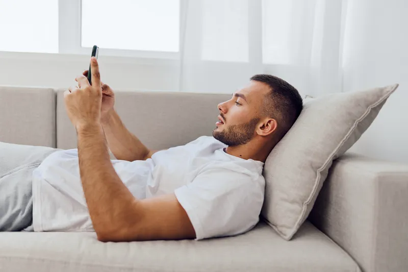 Relaxed man using smartphone while lying on a comfortable sofa, dressed casually in a white t-shirt. The modern interior creates a cozy atmosphere for leisure time.