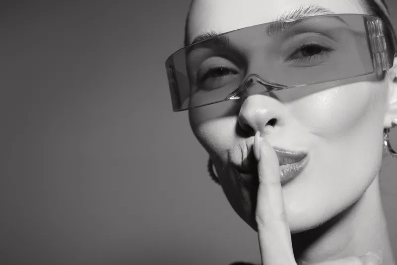 Close up of pale white woman with clear skin and transparent glasses making silence gesture on grey background.