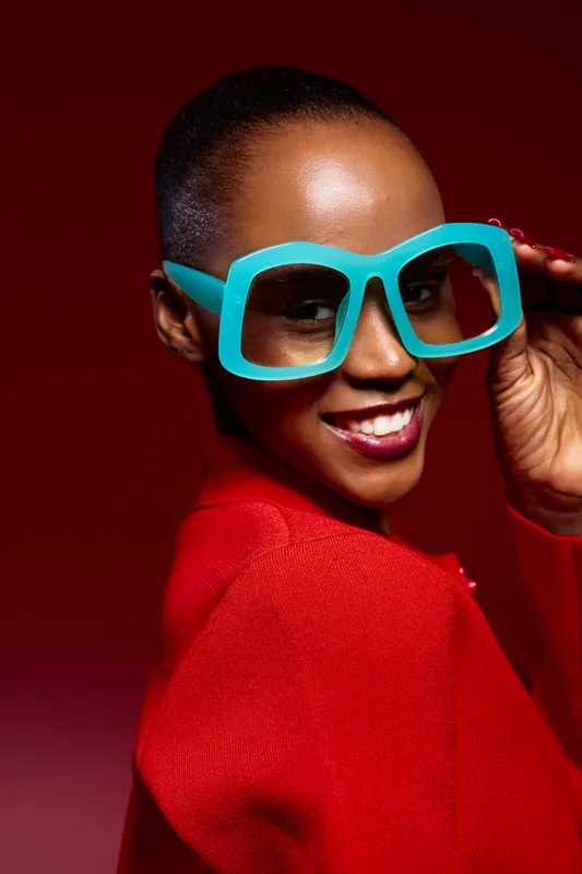 Close-up portrait of a smiling dark skinned African woman wearing oversized blue glasses and a red top. Bright studio lighting highlights her joyful expression and smooth skin tone.