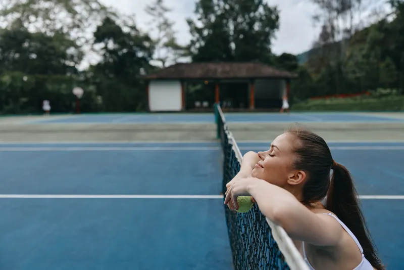 Young female tennis player resting by the court, enjoying a moment of relaxation in a serene outdoor setting, showcasing joy and determination