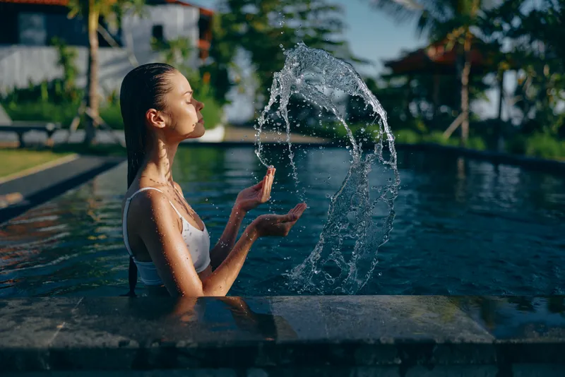 Young woman in white swimsuit enjoys outdoor pool lifestyle splashing water with hands under bright sunlight in tropical resort environment with blurred background.