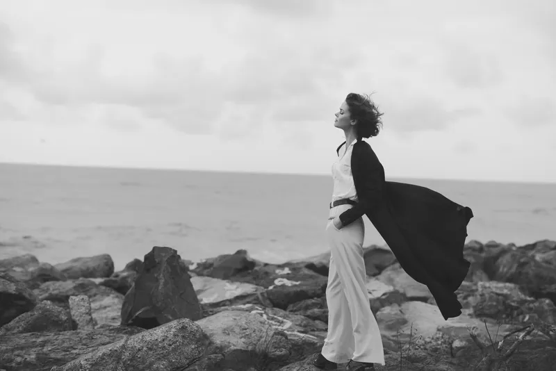 Wind sweeps along the coast as a cape billows behind a figure on rugged rocks, gazing toward the horizon, capturing solitude, strength, and maritime escape in monochrome.