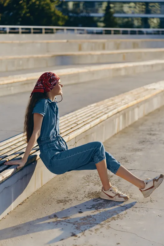 young woman sitting outdoors on concrete steps in casual denim jumpsuit with red headscarf during sunny day relaxing peacefully in urban park
