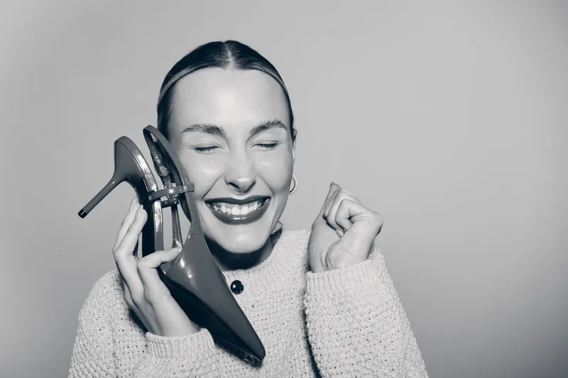 Happy Caucasian woman with fair skin holds high heel shoes near face, smiling with closed eyes in cozy sweater. Concept of joy and fashion.