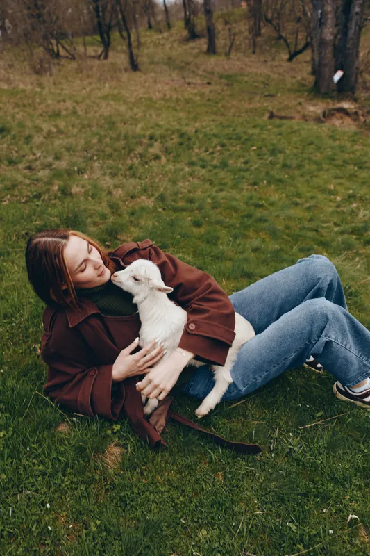 A young woman in a brown jacket lies on the grass holding a white baby goat close to her chest in a peaceful outdoor setting during autumn.