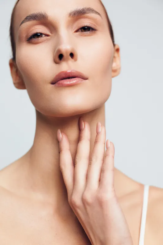Close-up portrait of a young woman with clear skin touching her neck gently, showcasing natural beauty and skincare concept with neutral background and soft lighting.