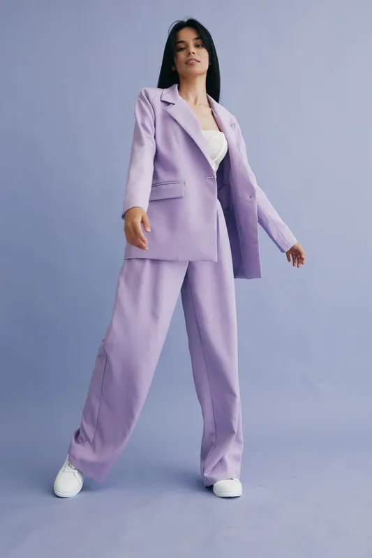Young woman wearing a lilac pantsuit and white sneakers stands confidently against a soft purple background, showcasing modern fashion and relaxed style.