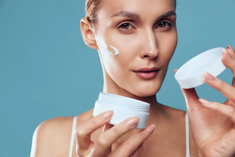 Close-up of woman applying white face cream on cheek, holding open jar with soft natural light. Concept of skincare, beauty routine, self-care, and healthy skin care.