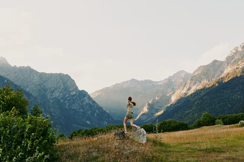 Cheerful woman outdoors, on a grassy hillside, distant mountains visible, relaxed pose, sunlit scene, soft breeze, vibrant natural colors