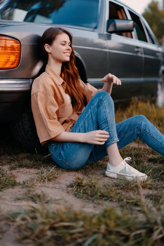Young woman sitting beside vintage car in casual outfit, enjoying a sunny day outdoors, expressing relaxed emotions.