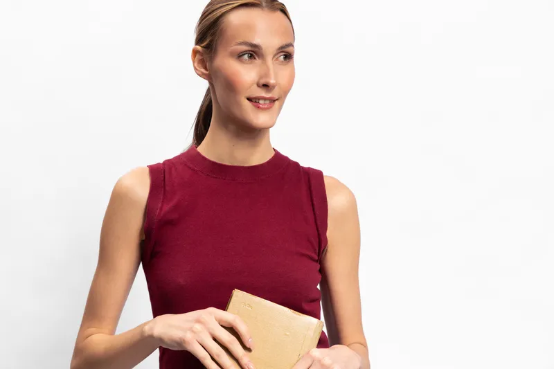 Young woman in sleeveless red top holds beige clutch purse, smiling slightly. Studio portrait on white background, fashion concept for casual style and accessories.