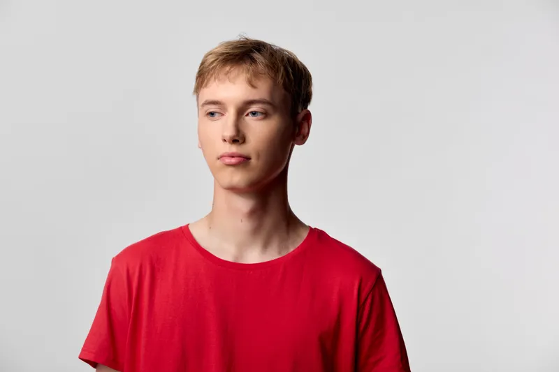 Young man in a red shirt stands against a neutral studio background with a thoughtful expression, casual style and clean pose suitable for fashion and lifestyle use
