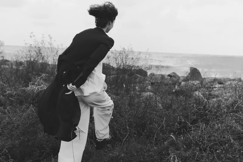 Hiker advancing through wind swept coastal grassland with backpack, exploring rugged shoreline and moody scenery in monochrome