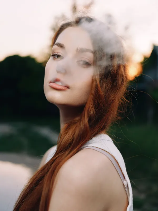 Young woman with long red hair exhaling smoke at sunrise in a natural setting, showcasing serene emotions and soft colors.