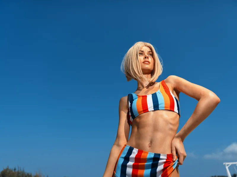 confident young woman wearing colorful striped bikini standing outdoors against clear blue sky, looking away with hand on hip, summer beachwear style