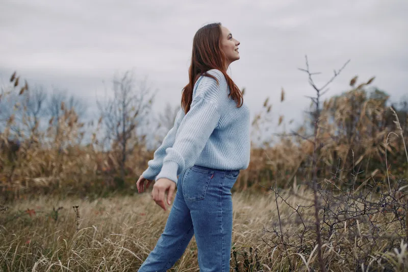 A candid, authentic moment of a woman in a cozy blue knit sweater and jeans walking through tall grass, capturing natural posture and genuine mood.