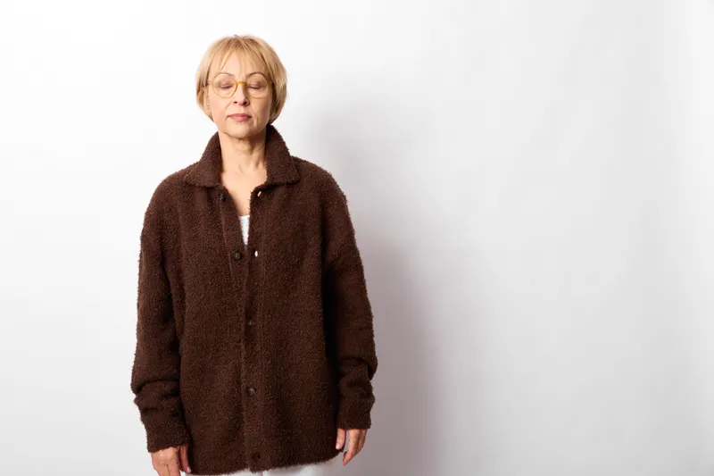 Elderly woman portrait standing in dark brown jacket and glasses, calm neutral expression, looking at camera, white studio background, minimal composition