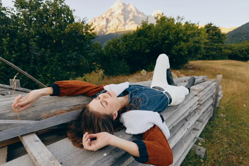 Woman relaxation on wooden platform in countryside meadow with mountain and nature views, lying outdoors in denim jacket and shearling, peaceful summer rest and scenic escape.