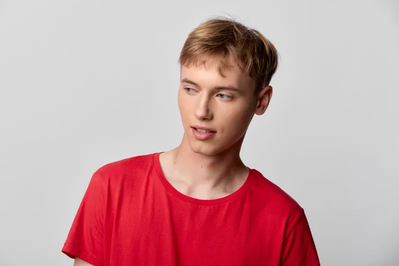 Youth, man in a casual red shirt looks away with a thoughtful expression, bright studio lighting against a clean background, contemporary everyday fashion vibe
