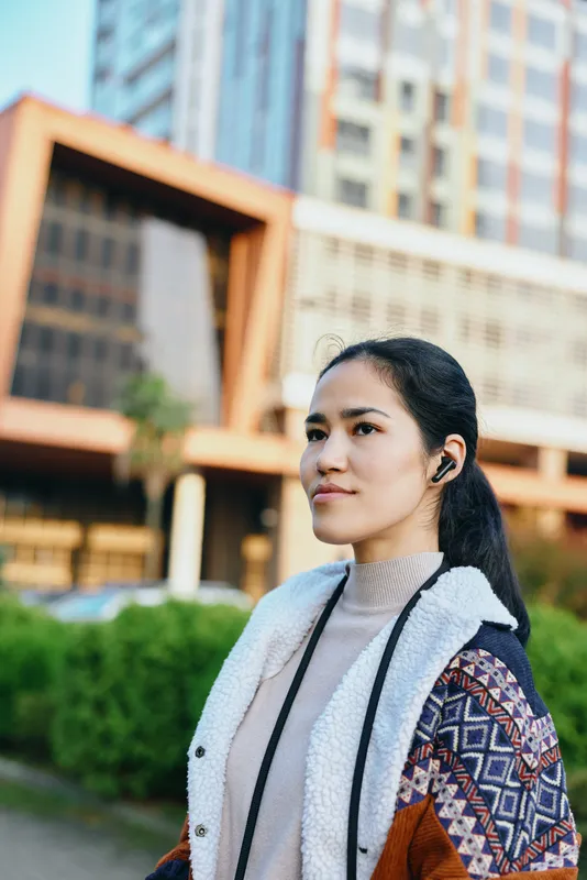 Woman in a stylish jacket and scarf stands outdoors near modern buildings, listening to wireless headphones in a calm urban setting with casual fashion