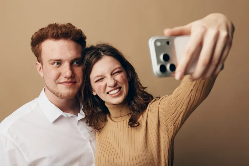 Young couple takes a selfie together, smiling and enjoying the moment. The woman wears a brown sweater, the man a white shirt, with a neutral beige background. Casual lifestyle concept.