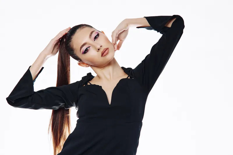 Young woman with dramatic makeup and long ponytail poses confidently in a stylish black outfit. Studio shot on white background for beauty, fashion, creativity concept.