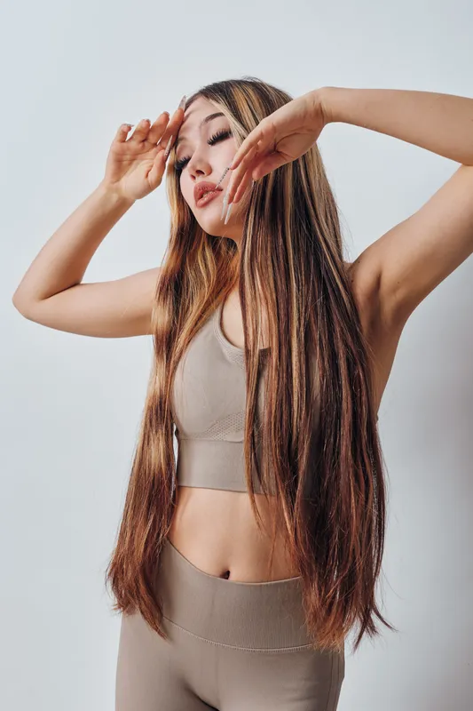 Woman with long braided hair wearing beige sportswear posing with eyes closed and hands near face in studio, showing relaxed and graceful posture for beauty concept.