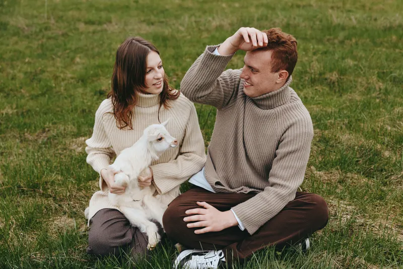 Couple sitting on grass with a white goat kid, wearing sweaters, enjoying a relaxed outdoor lifestyle moment with nature, affection, and gentle companionship in warm lighting, cozy ambiance.