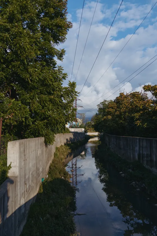 Urban canal with concrete canal walls and trees, water reflection beneath overhead power lines and cloudy sky; photographer shadow provides scale in this quiet drainage channel scene.