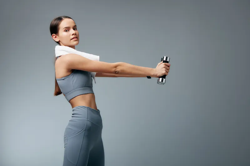 Fitness-focused woman in athletic wear extends arms with a dumbbell, towel around neck, studio backdrop, capturing strength, balance and determination during a dynamic workout session.
