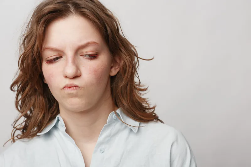Young woman with wavy hair looks confused and skeptical, wearing a light shirt against a plain background. Concept of doubt, uncertainty, and contemplation in emotions.